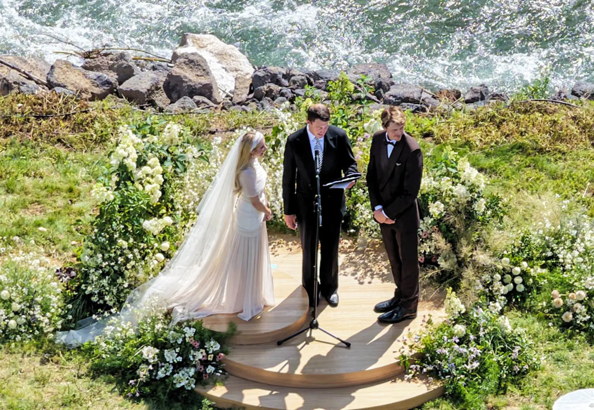 Emma Roberts and Cody John stand with an officiant at their wedding ceremony overlooking a river.