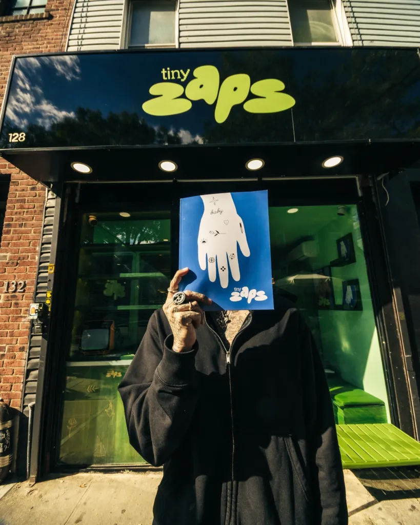 Person with tattooed arms holding a blue design booklet in front of their face, standing outside the Tiny Zaps tattoo shop.