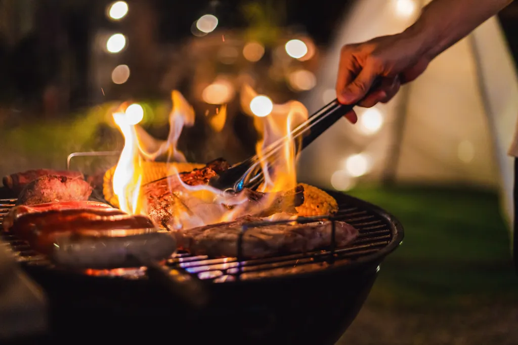 A person uses tongs to flip food on a flaming barbecue grill at night.