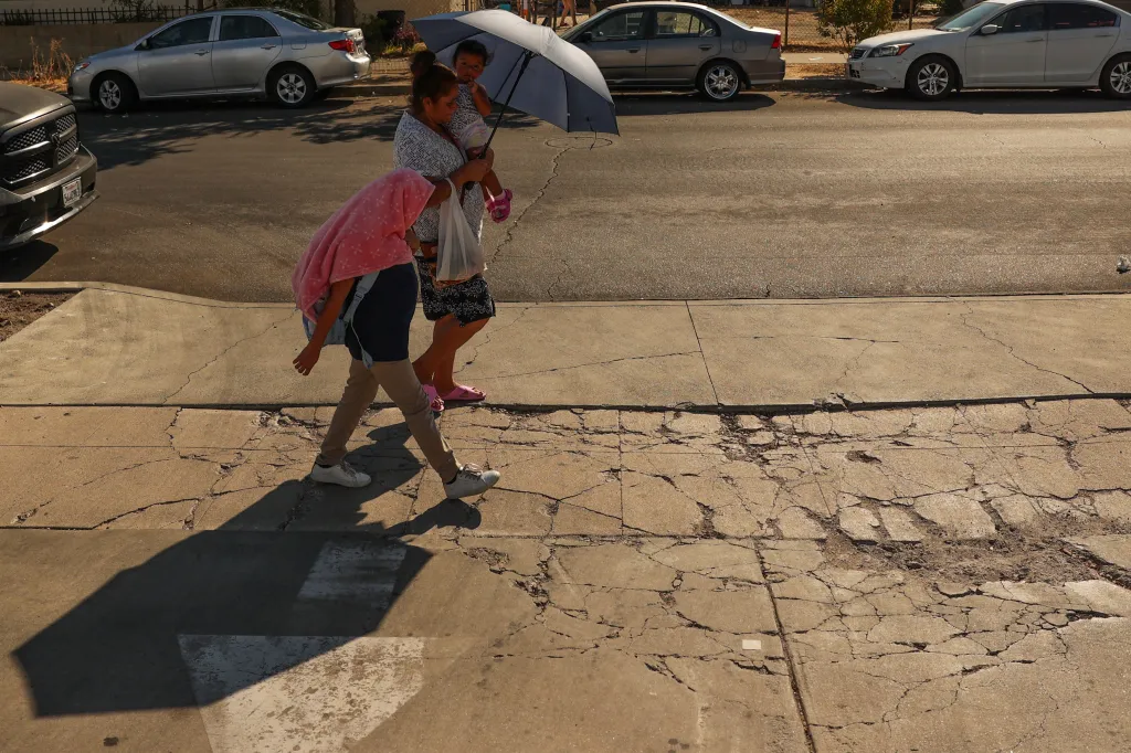 A family walks on a sidewalk in Pacoima, CA with a woman holding a child and an umbrella, and another child covering their head with a pink shirt due to hot temperatures.