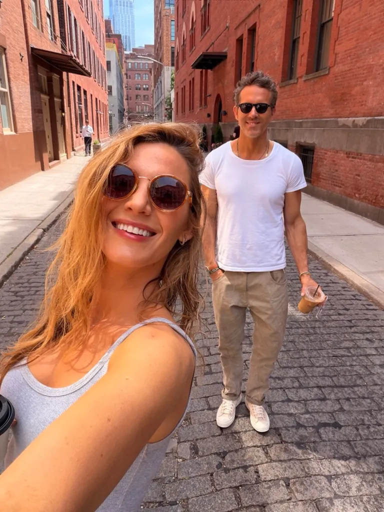 Blake Lively taking a selfie with Ryan Reynolds in the background on a cobblestone street.