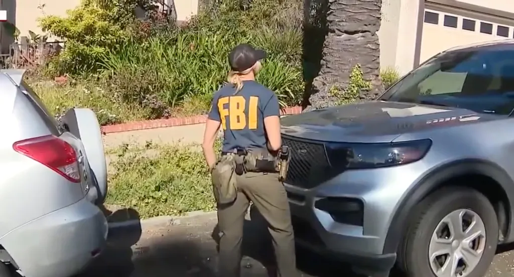 An FBI officer in a dark blue shirt with "FBI" in yellow stands next to a silver SUV.