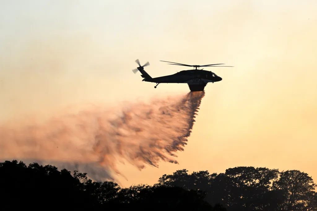 A fire helicopter drops water onto a wildfire in Sunol, California, silhouetted against the sunset.