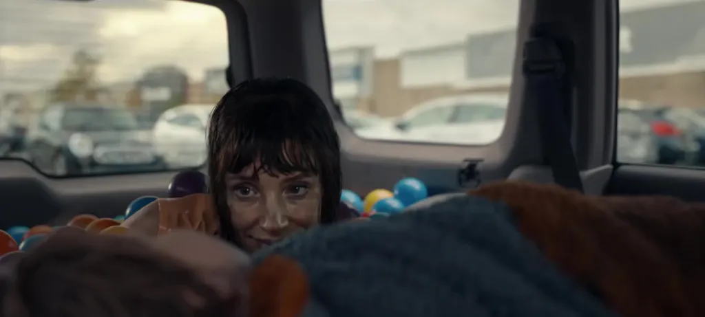 A woman with a creepy smile looks over the top of seats from a car trunk filled with colorful balls.