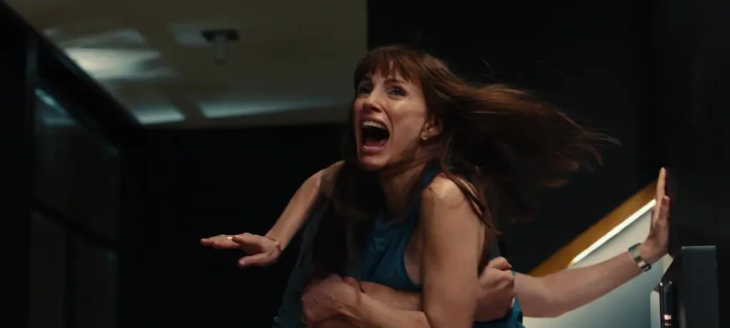 A woman with brown hair and bangs is screaming, wearing a blue sleeveless top, as a man's arms hold her from behind.