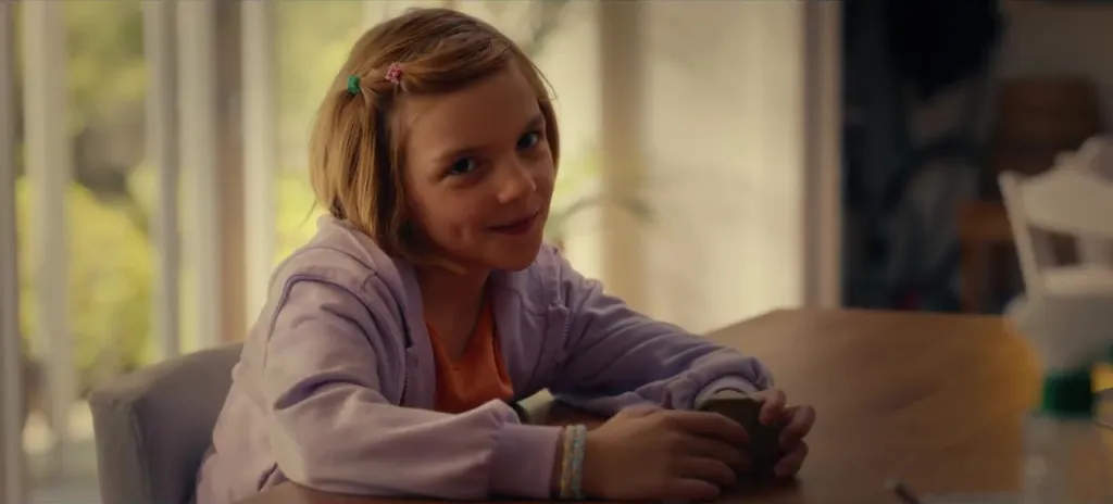 A young girl with blonde hair in pigtails smiles while sitting at a table.