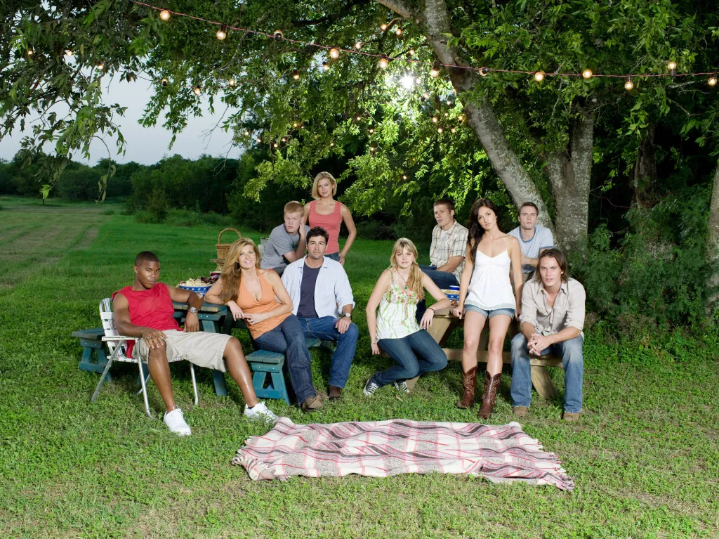 Cast portrait of the main cast of Friday Night Lights, including Gaius Charles, Connie Britton, Jesse Plemons, Adrianne Palicki, Kyle Chandler, Aimee Teegarden, Zach Gilford, Minka Kelly, Scott Porter, and Taylor Kitsch, gathered for a picnic under string lights.