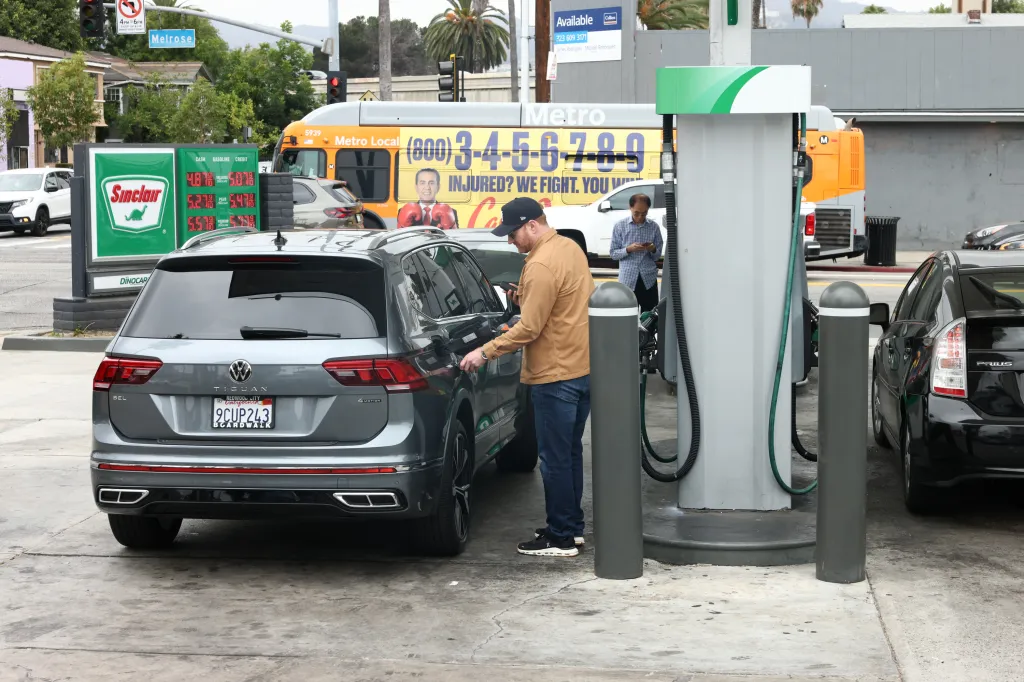 Man filling up a gray Volkswagen Tiguan at a Sinclair gas station where gasoline prices range from $4.87 to $5.57.
