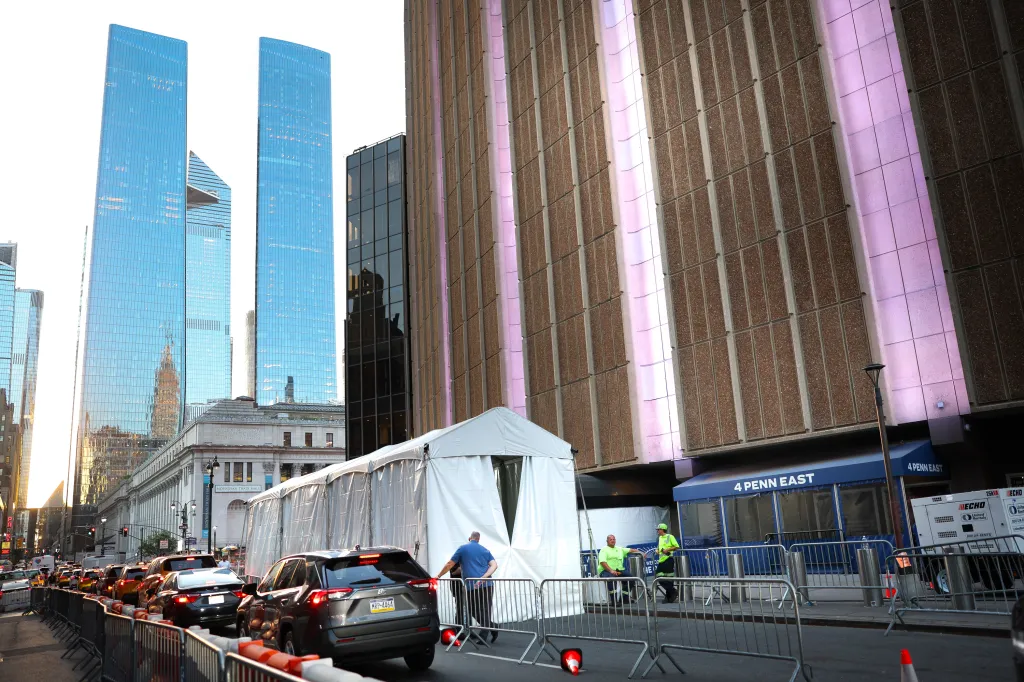 A long white tent and metal barriers block part of a city street in front of a building with glowing pink strips, with modern glass skyscrapers and an older building with columns visible behind.