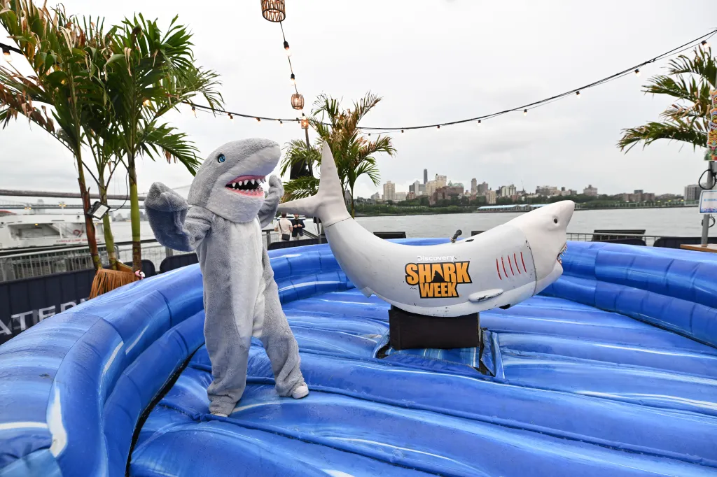 A person in a shark costume stands next to a mechanical shark ride at a Shark Week launch party.
