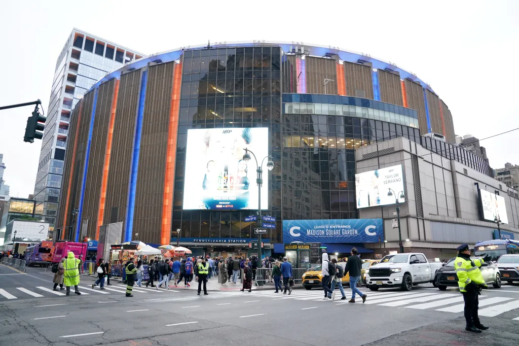 Exterior view of Madison Square Garden lit in orange and blue for the New York Knicks, with people crossing the street and traffic.