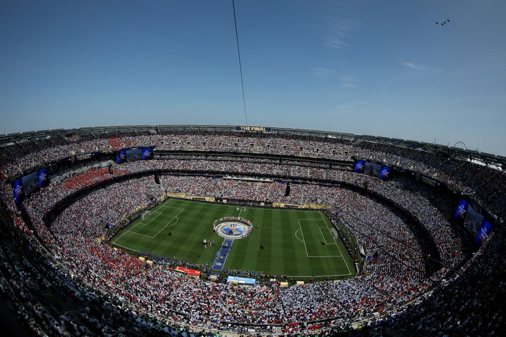 A general view of a soccer stadium packed with fans, with a large "FIFA World Cup 2026 THE FINAL" logo on the field and four F-16 fighter jets flying overhead.