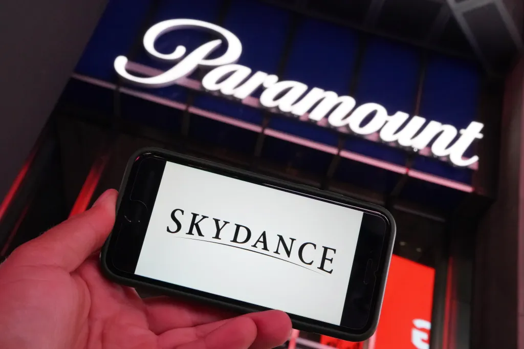 A hand holding a cell phone displaying the Skydance logo, with a blurred Paramount sign in the background.