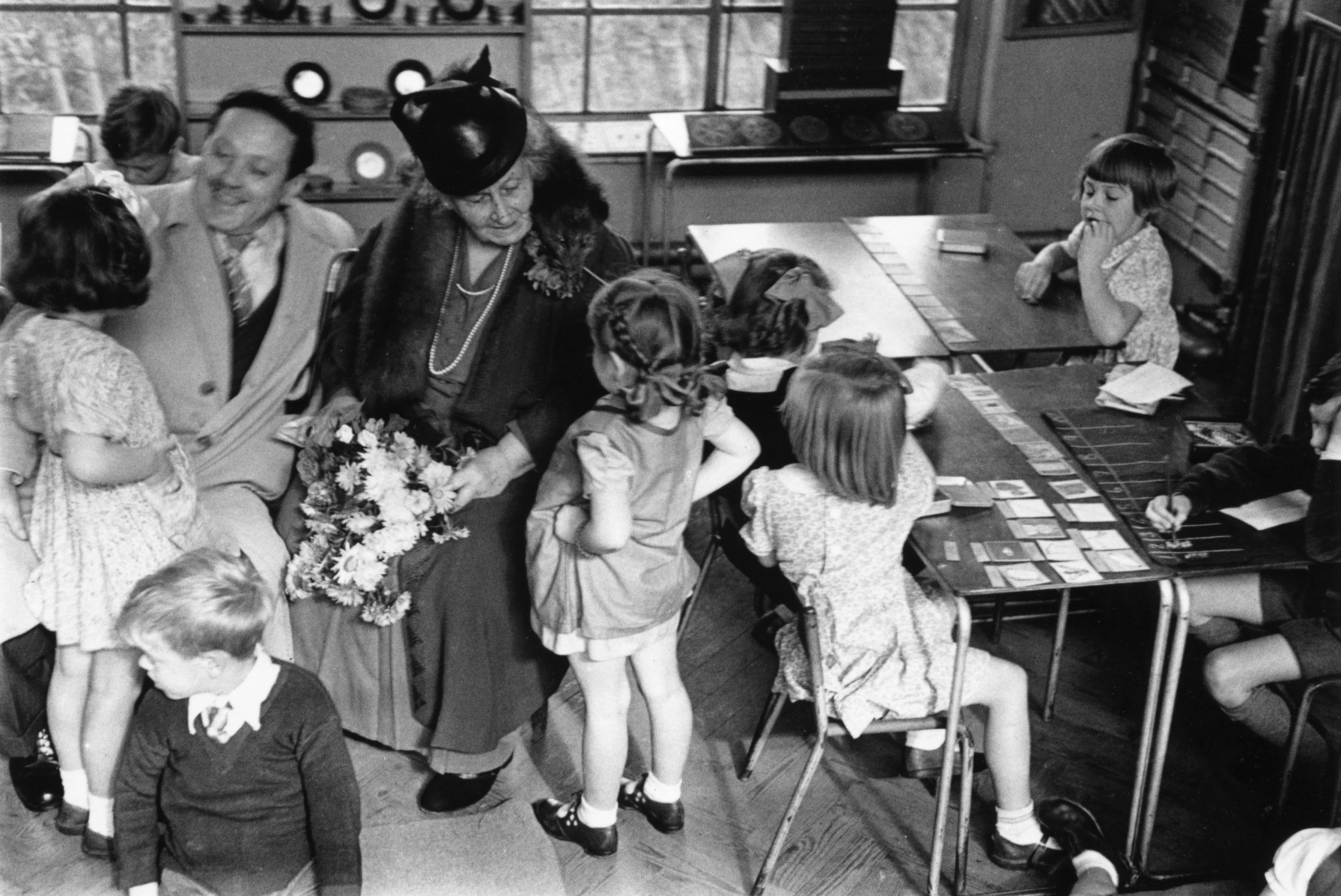 Maria Montessori sitting with a group of children learning with hands-on objects in the 1940s.