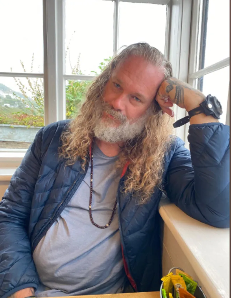 A man with long curly hair and a gray beard, wearing a blue puffer jacket, looks towards the camera.