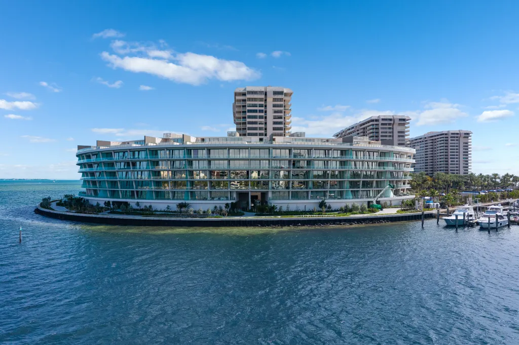 A modern, curved building with glass balconies overlooking the water, with older apartment buildings and a marina in the background.