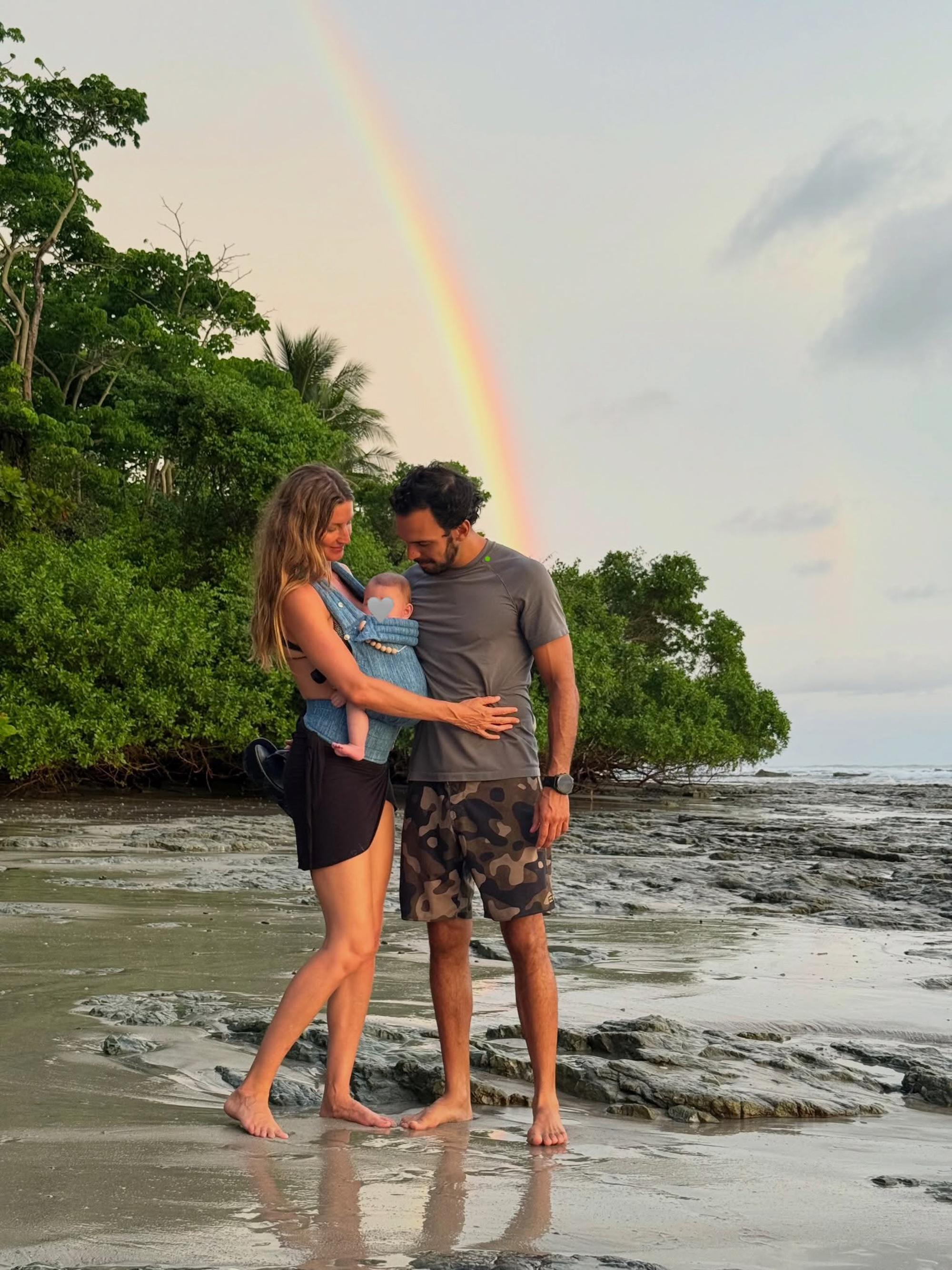 Gisele Bündchen and Joaquim Valente stand on a beach, holding a baby in a carrier, with a rainbow visible in the sky.