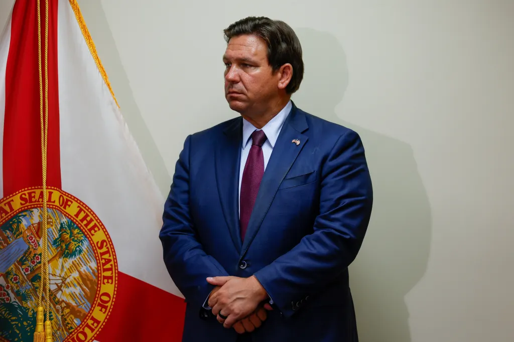 Florida Gov. Ron DeSantis in a navy suit and red tie stands next to the Florida state flag.