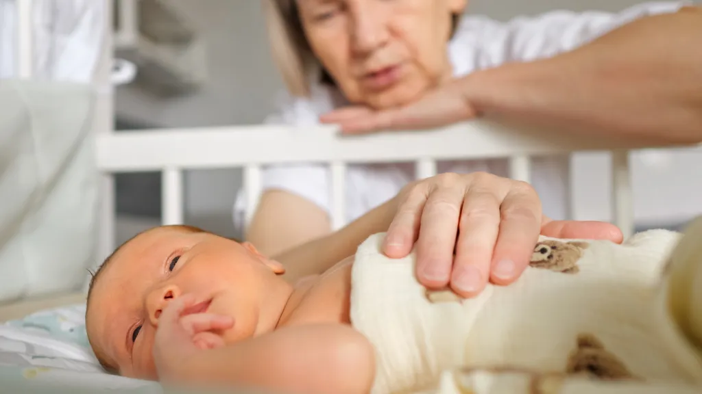 A grandmother gently touches a newborn baby swaddled in a crib.