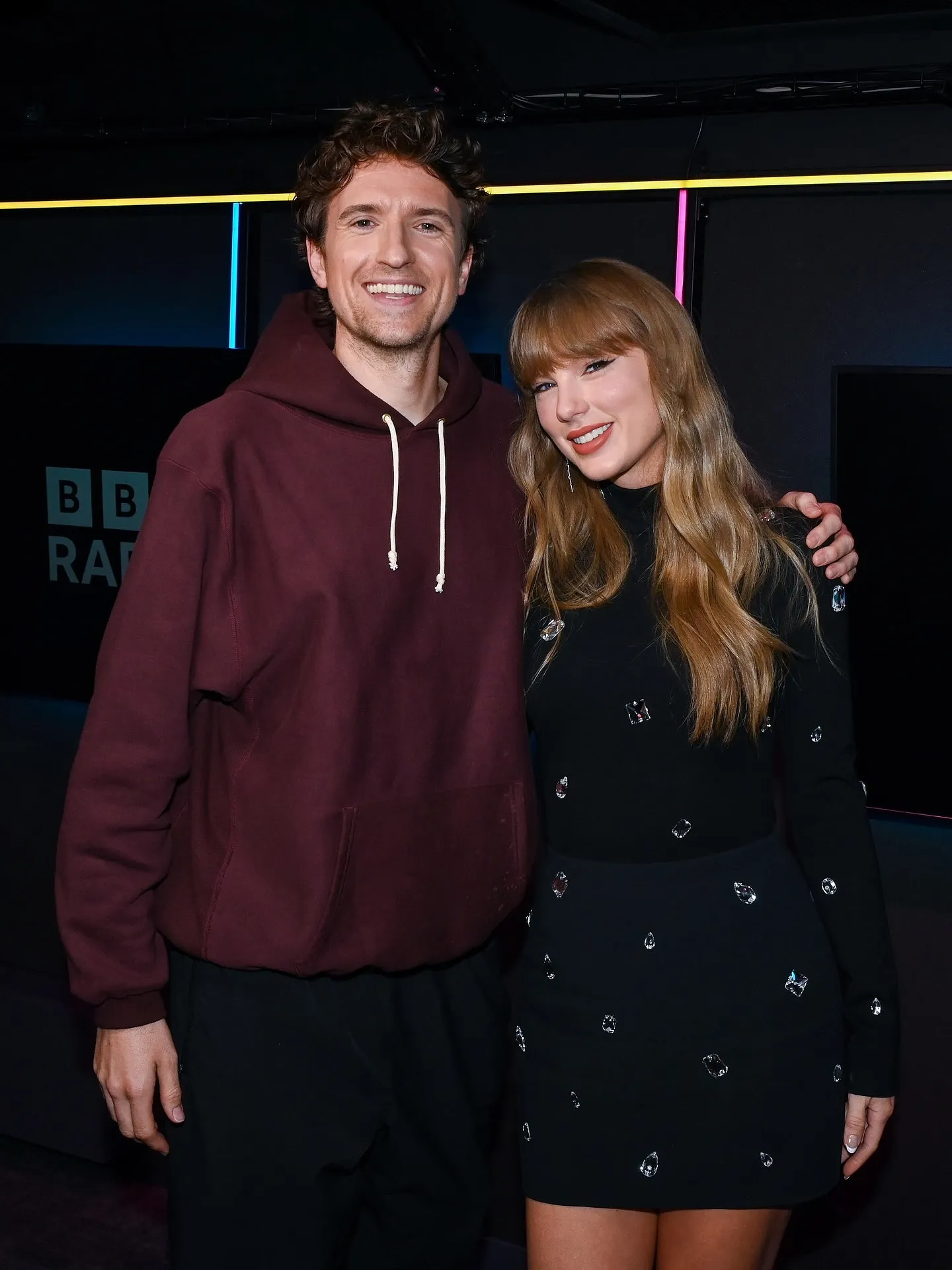 Greg James and Taylor Swift standing together, smiling, with James's arm around Swift.