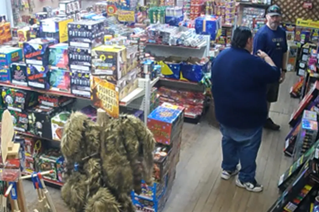 Two men in a fireworks store, the one in the foreground wearing a blue shirt and jeans, and the other man in a blue shirt and baseball cap.