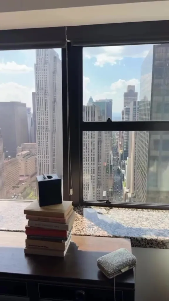 View from a high-rise window overlooking a city street and buildings, with a stack of books, a tissue box, and a sparkly clutch purse on a table in the foreground.
