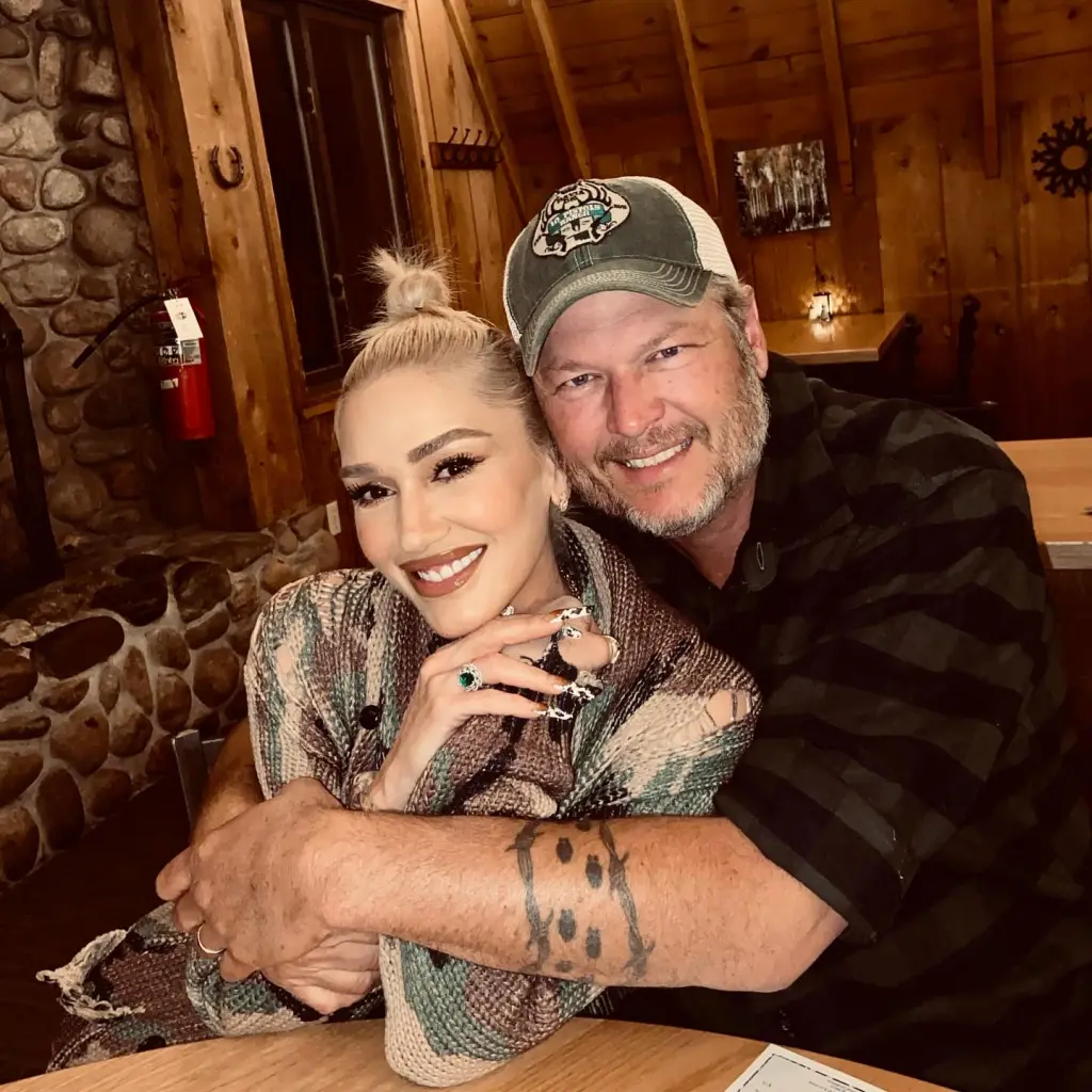 Gwen Stefani and Blake Shelton smiling at the camera, with Blake Shelton's arm wrapped around Gwen Stefani.