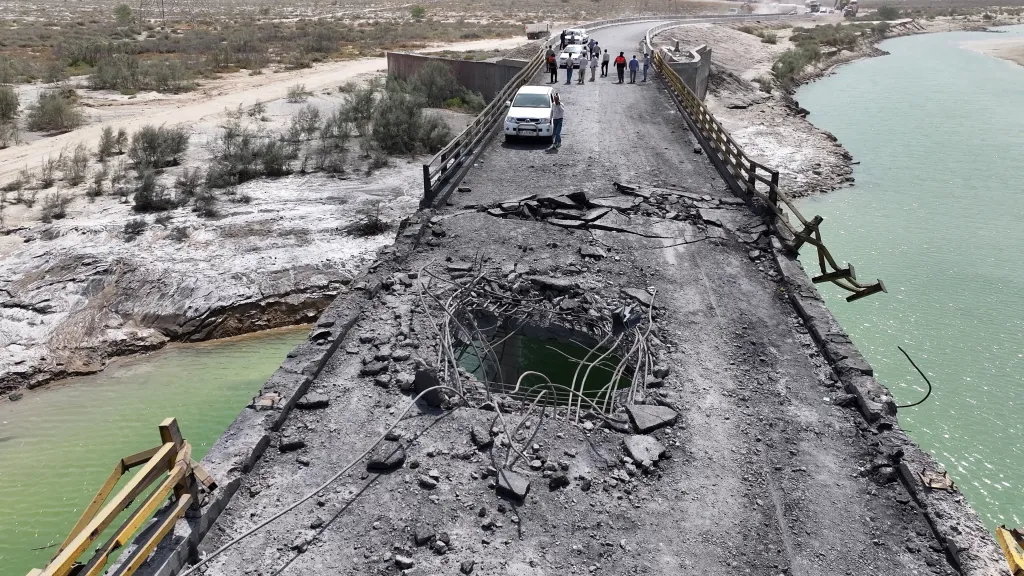 A large hole on a damaged bridge, with a green river flowing below, and people and a white car on the intact section of the bridge.