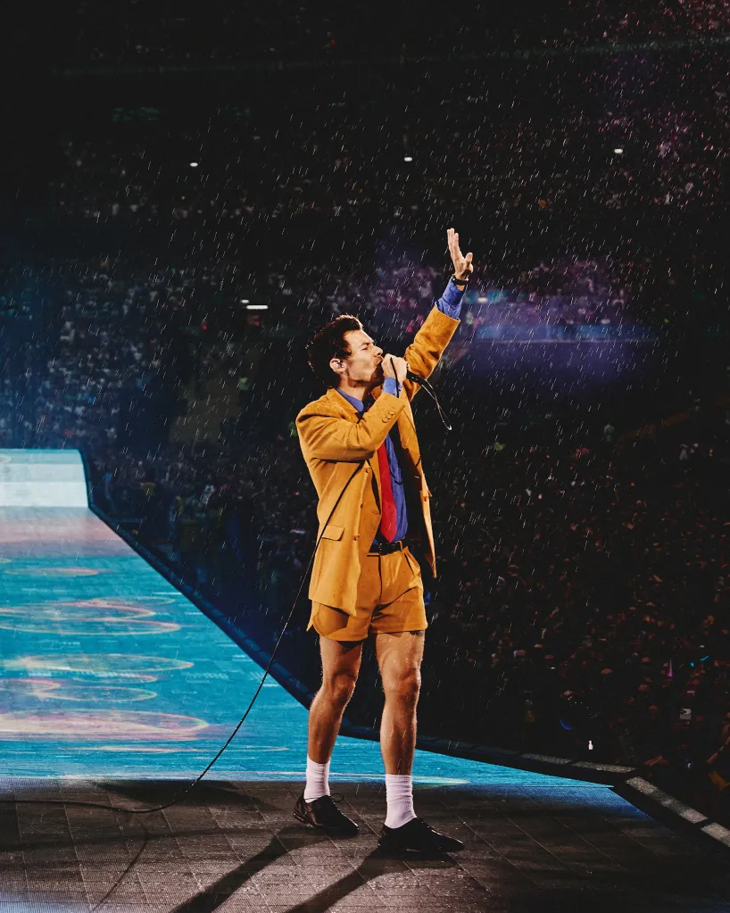 Harry Styles in concert, wearing a gold suit and holding a microphone, with a crowd and stage lights in the background.