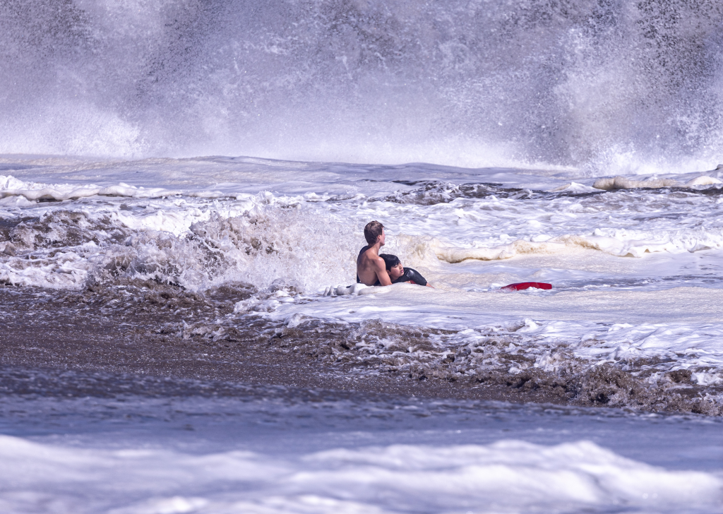 A young lifeguard holding a boy in a wetsuit, both submerged in rough ocean waves with a red boogie board nearby.