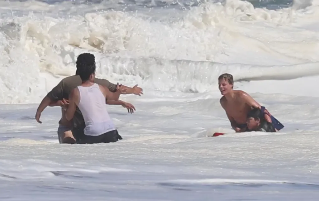 A young lifeguard carrying a child through foamy water while two adults stand nearby in the rough ocean waves.