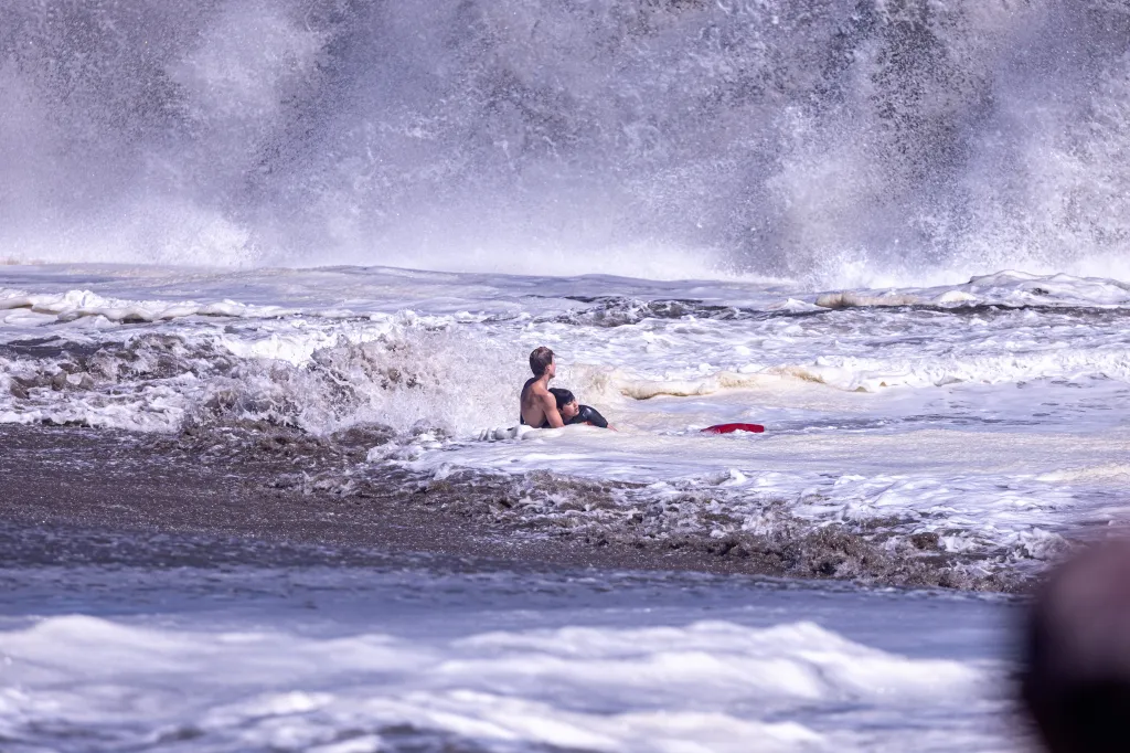 A boy in the water, wearing a black top, is battered by large waves, with a red bodyboard nearby.
