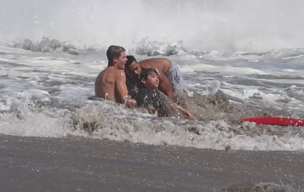 Three people, two adults and one child, in ocean waves with a red rescue board partially visible.