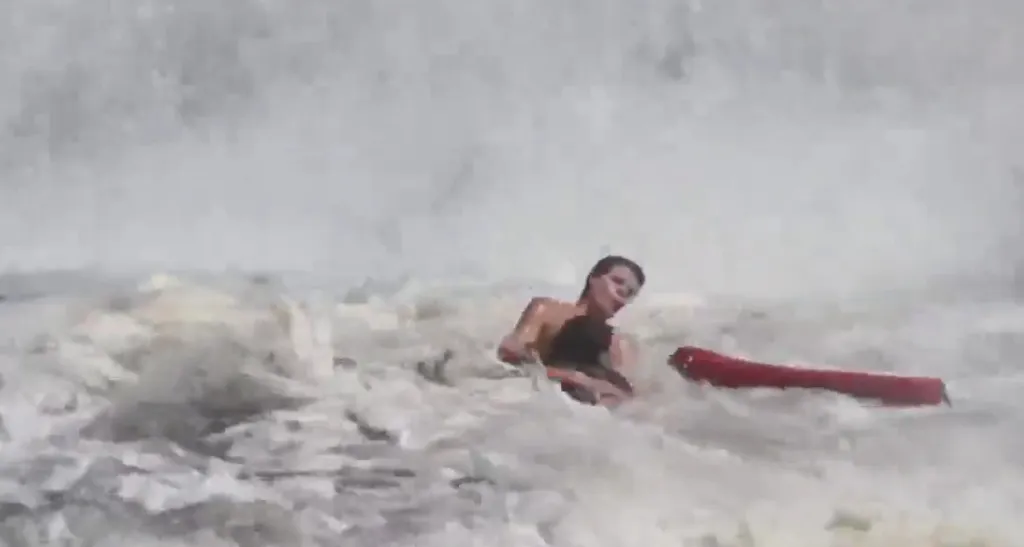 A young lifeguard rescuing a boy from huge waves with a red rescue tube.