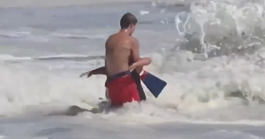 A lifeguard in red shorts carrying a small boy from the ocean, as waves crash around them.