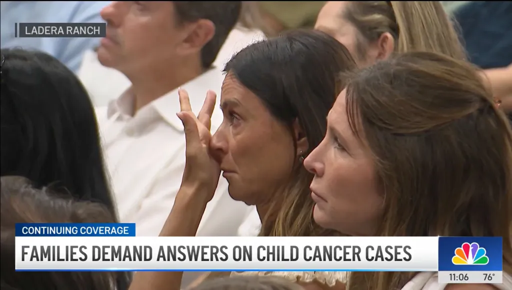 A woman wipes tears from her face as other people in the audience look on, with a news chyron at the bottom reading "Families Demand Answers on Child Cancer Cases".