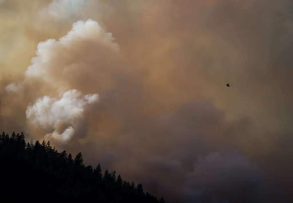 A helicopter flies above a dark tree line with heavy smoke and dark clouds in the background.