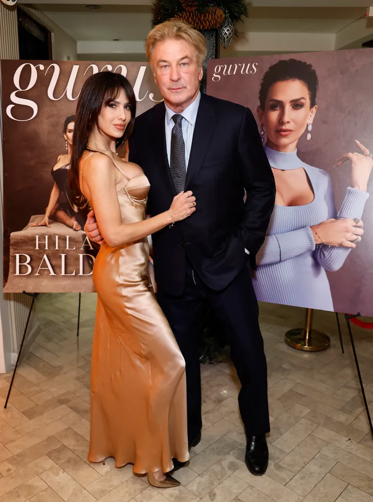 Hilaria Baldwin in a satin dress and Alec Baldwin in a suit stand in front of two posters featuring Hilaria Baldwin on the cover of Gurus Magazine.