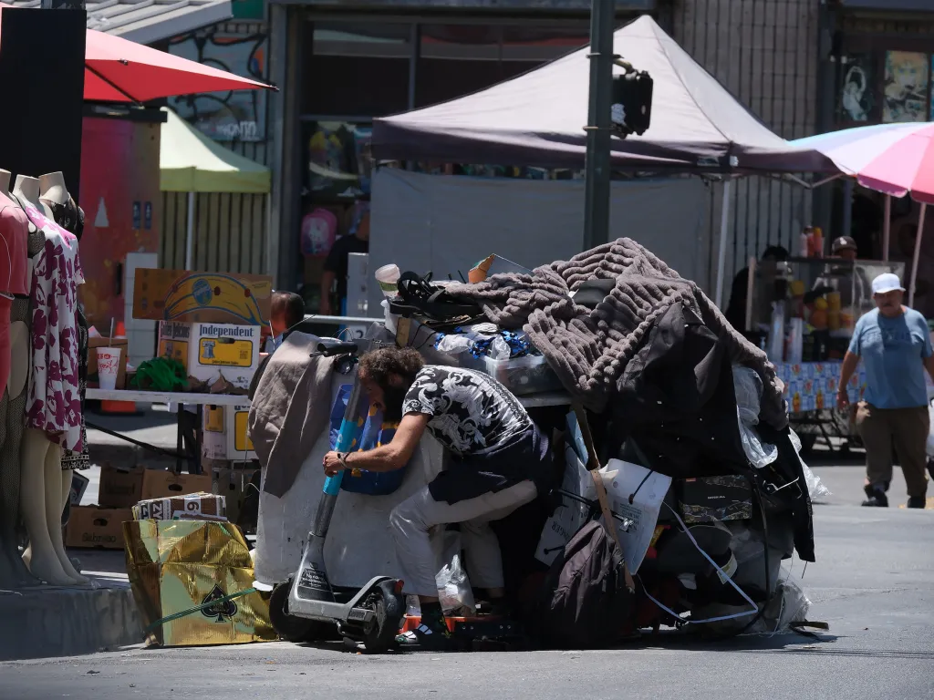 A person bent over, pushing a scooter laden with belongings and blankets.