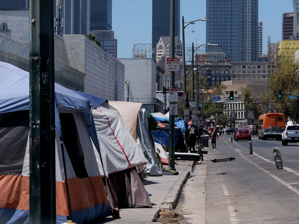 Tents and tarps line a street in downtown Los Angeles, with city buildings visible in the background.