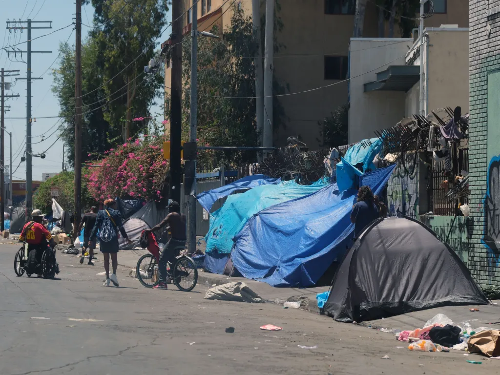 A street scene in Los Angeles with several homeless individuals, tents, and blue tarps along the sidewalk, and trash on the ground.
