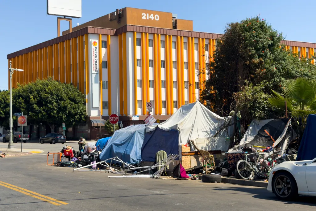Tents and belongings of homeless people line Olympic Boulevard in Los Angeles, contrasting with the Koreatown ADHC building in the background.