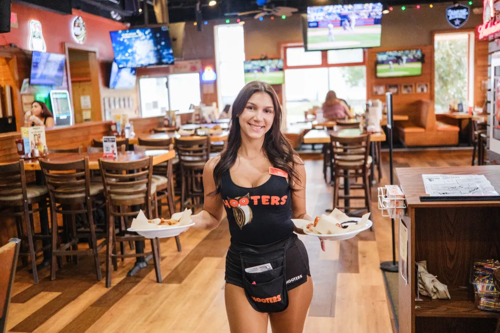 Hooters server Sofia Nizzo holding two plates of wings inside a Hooters restaurant.