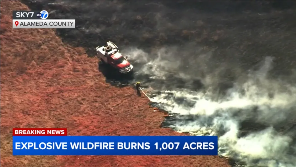 Aerial view of a wildfire burning 1,007 acres in Alameda County, with a red fire truck and a firefighter battling the blaze.