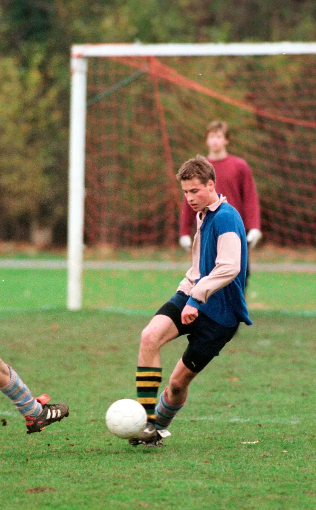 Prince William in his school's fawn and blue colors, kicking a soccer ball during a game.