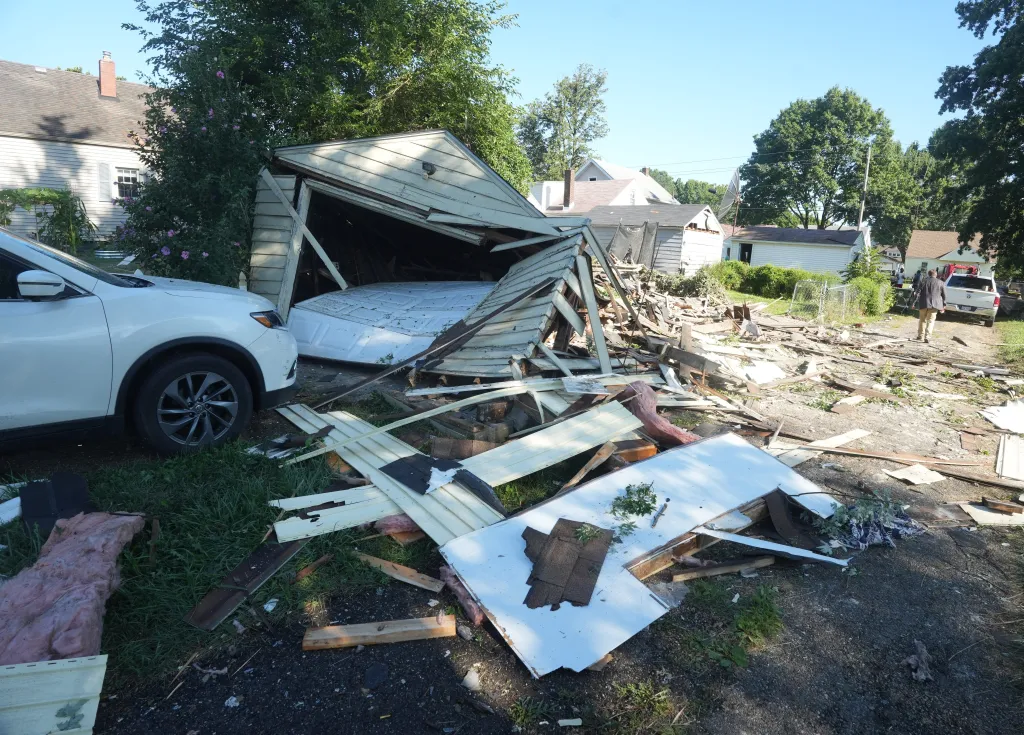 Debris from an exploded house scattered across a neighborhood, damaging a white car and a garage, with people surveying the scene in the background.