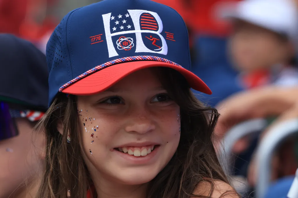 A young girl with long brown hair, wearing a red, white, and blue baseball cap and red, white, and blue glitter on her face, smiles while looking to the side.