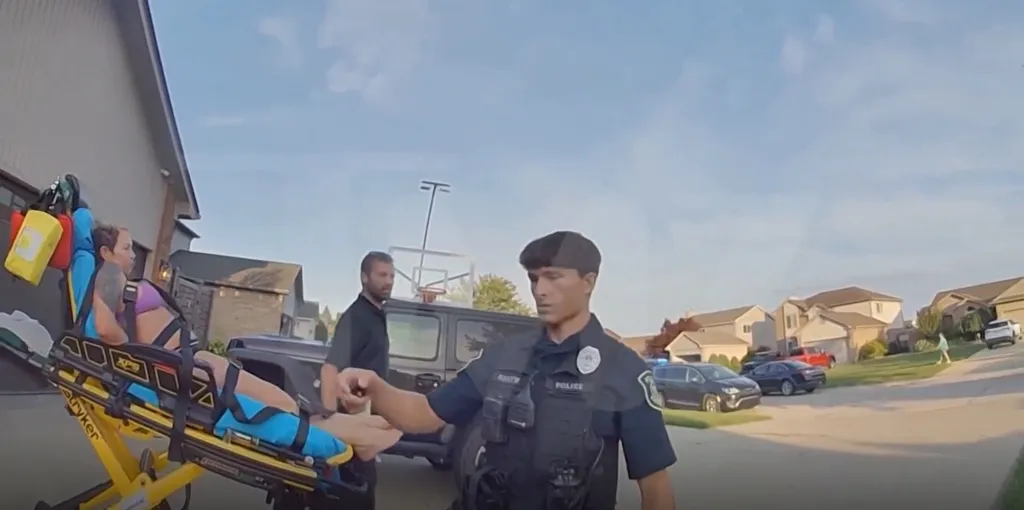 A police officer and another man stand near a woman in a purple bra and black bottoms who is strapped to a stretcher outside a house.