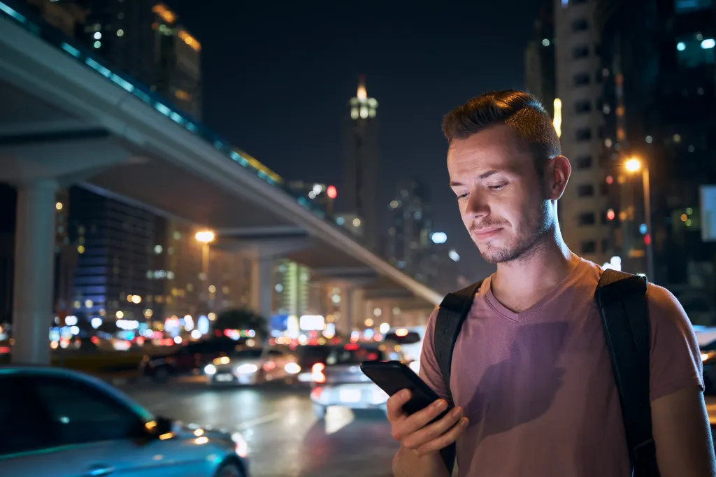 A man with a backpack using his phone on a busy city street at night.