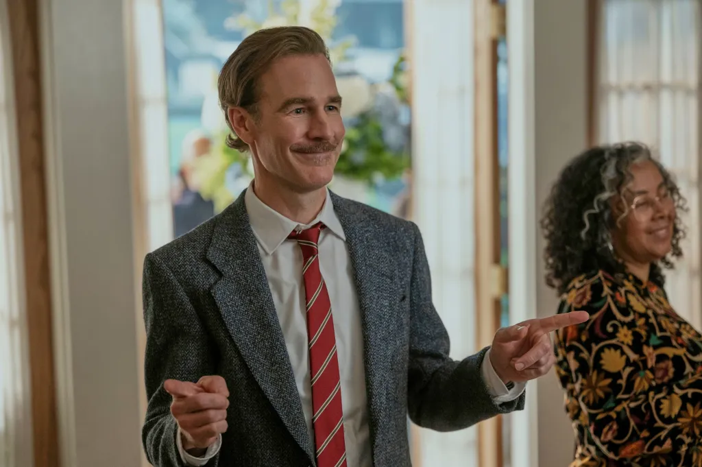 James Van Der Beek as a character with a mustache and a patterned suit, pointing with both hands, with another person in the background.